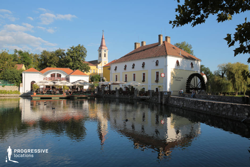 This location features a historic watermill building with a large wooden waterwheel positioned alongside a calm lake. The architecture is characterized by a traditional European style, with a yellow plaster facade, red-tiled roof, and decorative window accents. The structure appears well-maintained and blends harmoniously with the natural surroundings. Adjacent to the watermill, a lakeside restaurant with an open terrace extends over the water, offering an inviting setting with outdoor seating under large parasols. The reflection of the buildings and trees in the still water enhances the tranquil atmosphere. A stone walkway with a low stone wall runs along the lake, providing access to the area and integrating seamlessly with the historic aesthetic. In the background, a church with a sharp, red spire and classical architectural details rises above the surrounding structures, contributing to the picturesque small-town ambiance. Lush greenery, including mature trees and willows drooping over the water, adds to the scenic and serene quality of the location. This setting is ideal for capturing idyllic European townscapes, historical architectural features, and peaceful waterfront scenes. The blend of built heritage and natural elements makes it a versatile filming or photography location suitable for period settings, travel content, or romantic scenes.