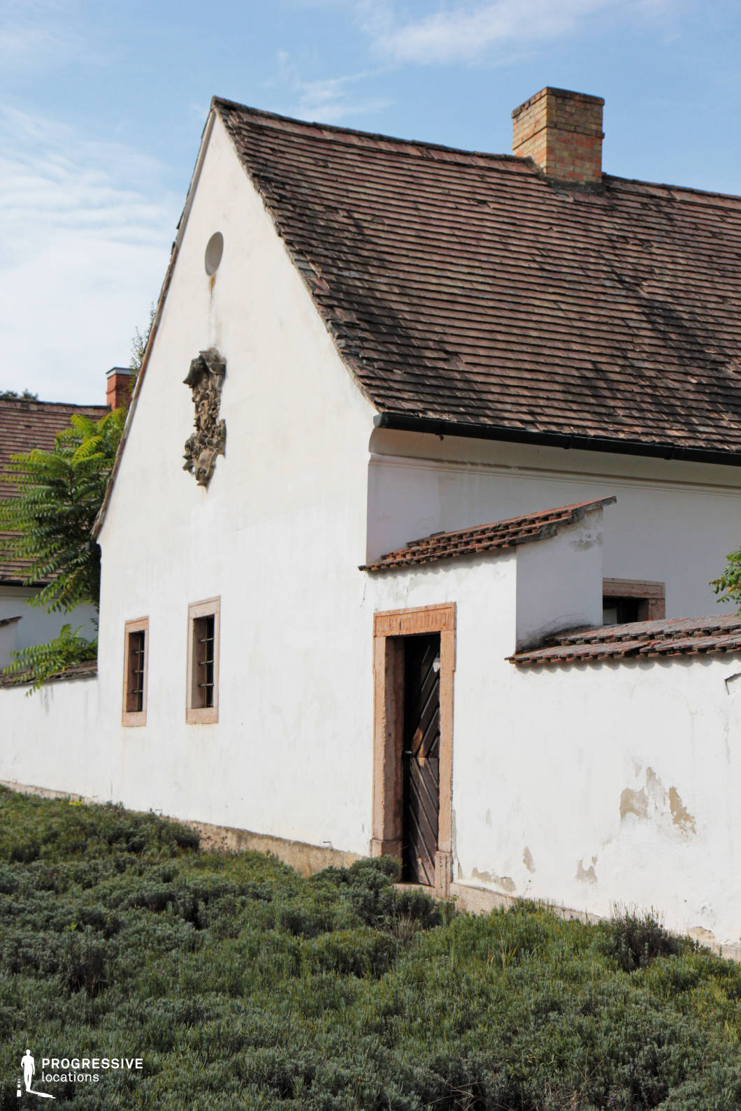 This location showcases a traditional European rural building with a weathered white plaster facade and a steeply pitched, aged tile roof. The structure has a simple yet authentic aesthetic, with exposed brickwork around the chimney and wooden-framed windows that enhance its historic character. A decorative relief adorns the upper gable, adding an element of craftsmanship. The building features a partially open wooden door set in a slightly eroded stone frame, leading into an enclosed courtyard. The surrounding low walls and adjoining structures contribute to the rustic and intimate feel of the space. The foreground is covered in dense, low-growing vegetation, which enhances the organic, unpolished charm of the location. The combination of aged textures, muted colors, and traditional architectural elements makes this setting ideal for period film productions, countryside-themed photography, or historical documentary visuals. The contrast between the well-preserved decorative elements and the natural wear of the structure adds visual depth and storytelling potential.