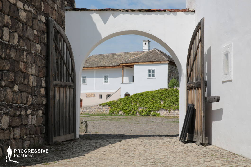 This image captures a rustic, historic stone gateway with an arched entrance, opening into a courtyard framed by a white-walled building. The wooden doors, aged and weathered, are set within thick stone walls, emphasizing a traditional and fortified architectural style. The cobblestone ground leads toward a secondary structure with a sloping tiled roof, white plastered walls, and wooden accents, evoking a Mediterranean or Central European rural aesthetic. The presence of a small chimney and ivy-covered stone retaining walls adds to the timeless charm of the scene. The composition creates depth and perspective, making it a compelling location for period film settings or heritage-inspired productions.