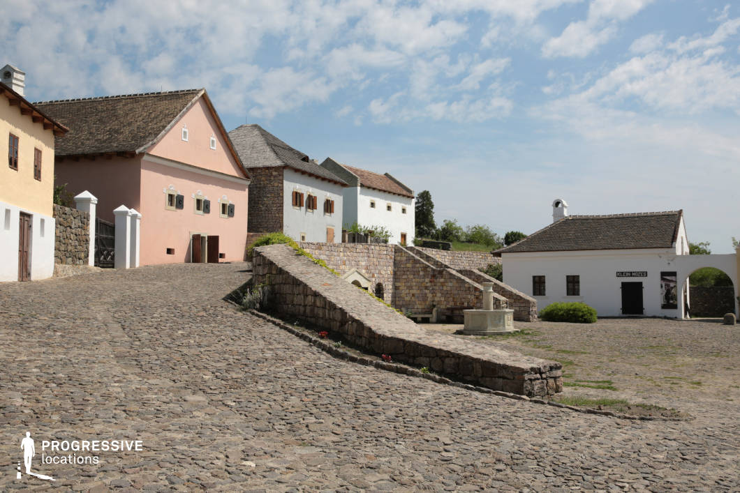 This image showcases a charming village square paved with irregular cobblestones, surrounded by traditional European-style buildings with distinct architectural features. The pastel-colored facades, wooden shutters, and sloping tiled roofs contribute to a rustic yet inviting atmosphere. A central stone retaining wall and staircase create a sense of depth and elevation, leading to upper-level structures that appear to be residences or historic buildings. The small fountain in the foreground adds a focal point, enhancing the authenticity of the scene. The white building with an arched entrance and signage suggests a former marketplace or artisan shop, reinforcing the village's historical character. This location is ideal for period film settings, heritage documentaries, or commercial projects seeking an old-world European ambiance.
