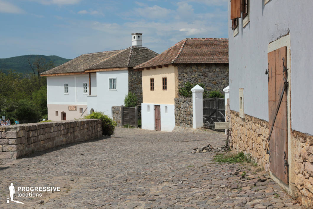 This image depicts a gently sloping cobblestone street lined with historic stone and plaster houses, evoking the ambiance of a traditional European rural village. The buildings showcase a blend of architectural styles, with exposed stone walls, pastel-colored facades, wooden shutters, and aged wooden doors reinforced with metal fittings. White-washed walls contrast with the natural stone elements, enhancing the rustic aesthetic. A stone retaining wall borders the path, leading toward a background of rolling green hills, adding a scenic depth to the setting. The wooden gates and enclosed courtyards suggest a preserved, heritage-rich environment. This location is well-suited for historical and period film productions, offering an authentic, old-world European charm.