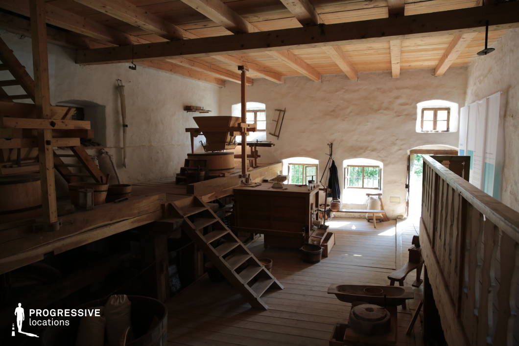 This image captures the interior of a historic mill workshop, featuring a rustic wooden structure with exposed beams and a hand-crafted wooden floor. The space is dimly lit, with natural light filtering through small, arched windows set into the thick, textured plaster walls, enhancing the old-world ambiance. A variety of traditional milling equipment, including wooden chutes, grinding mechanisms, and storage containers, are arranged throughout the workshop. A wooden mezzanine with a simple railing overlooks the workspace, connected by a sturdy wooden ladder. The presence of burlap sacks, wooden tools, and hand-carved details reinforces the authenticity of this heritage site, making it a suitable location for period films, historical documentaries, or educational projects focused on traditional craftsmanship.