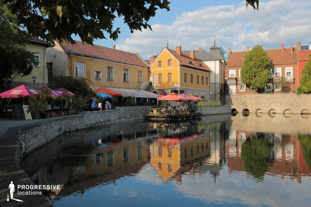 This image showcases a charming European old town with a scenic canal, where historic buildings in warm pastel tones line the waterfront. The architecture blends traditional elements such as red-tiled roofs, wooden shutters, and stone facades, creating a rich, inviting atmosphere. The canal’s still water reflects the colorful buildings and surrounding greenery, adding depth and symmetry to the composition. A stone embankment with arched openings reinforces the historical character of the setting. On the left, outdoor cafés with vibrant umbrellas and potted plants enhance the lively, social ambiance of the space. This location is ideal for film productions or commercial projects seeking a romantic, Mediterranean-style old-town aesthetic.