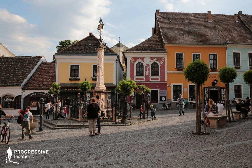 This image captures a picturesque European old town square, paved with cobblestones and surrounded by colorful historic buildings with ornate facades. The central monument, a tall stone column topped with a decorative cross, serves as a focal point, enclosed by a wrought-iron fence and accented by small, well-manicured trees. The buildings feature traditional architectural details, including intricate stucco reliefs, arched windows, and wooden shutters, painted in warm and inviting hues. The presence of street cafés and small businesses reinforces the lively, communal atmosphere of the square. This location is ideal for film productions, travel documentaries, or commercial projects seeking an authentic European old-town ambiance with a mix of history and vibrant street life.