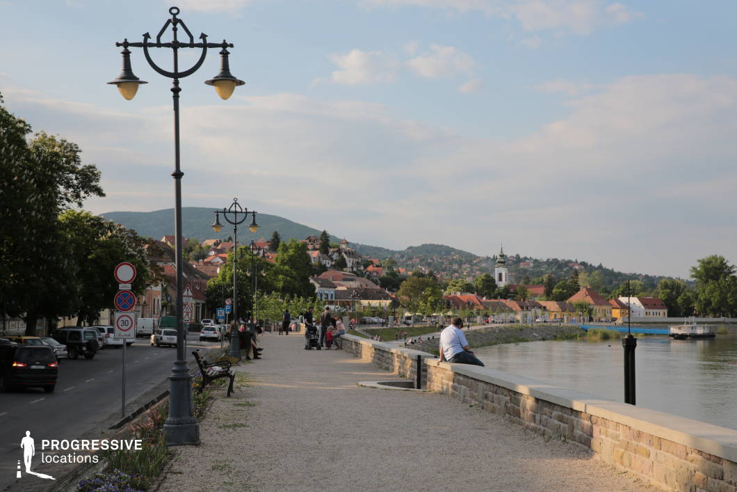 This image captures a charming riverside promenade lined with elegant wrought-iron lampposts, stone benches, and well-maintained walkways. The path runs parallel to a calm river, separated by a low stone wall, offering an inviting space for relaxation and social interaction. In the background, a picturesque townscape unfolds, featuring red-tiled rooftops, colorful facades, and a distinctive church tower, all nestled among lush greenery and rolling hills. The mix of historic and modern elements, combined with the waterfront setting, creates a serene yet lively atmosphere. This location is ideal for film productions, travel documentaries, or commercial projects that require an authentic European riverside town ambiance.