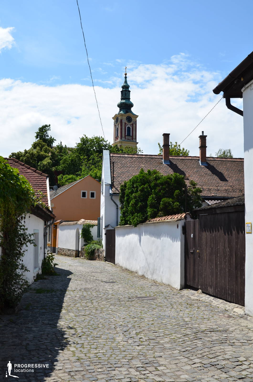 This image captures a serene cobblestone street in a European village, lined with white-washed walls and traditional tiled-roof houses. The winding pathway leads towards a striking Baroque-style church tower with an ornate clock face and green spire, rising above the rooftops. The architecture features rustic wooden gates, arched doorways, and lush greenery creeping along the walls, adding to the old-world charm. The scene conveys a peaceful, timeless ambiance, making it an ideal location for period film productions, travel documentaries, or commercial projects that seek to capture the essence of a picturesque European village.