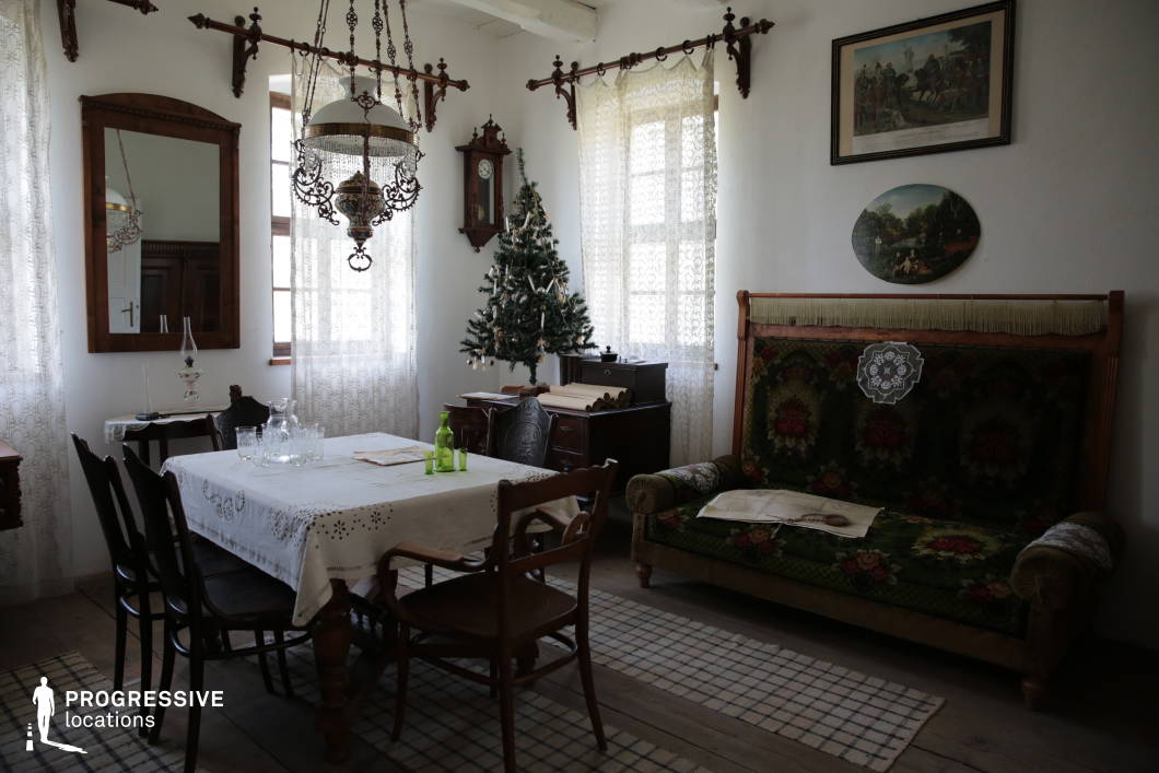 This image captures the interior of a traditional European village home, featuring vintage furniture and ornate decor. The room is adorned with antique wooden furnishings, including a carved dining table with a lace tablecloth, black wooden chairs, and an upholstered wooden-framed sofa with intricate floral patterns. The space is illuminated by natural light filtering through delicate lace curtains, which soften the atmosphere and highlight the period details. A decorative chandelier with intricate metalwork hangs from the ceiling, while an old-fashioned wall clock and framed historical artworks add to the nostalgic charm. The wooden flooring and area rugs contribute to the cozy, lived-in feel. This setting is ideal for historical reenactments, period films, or heritage-focused projects capturing the essence of traditional European rural life.