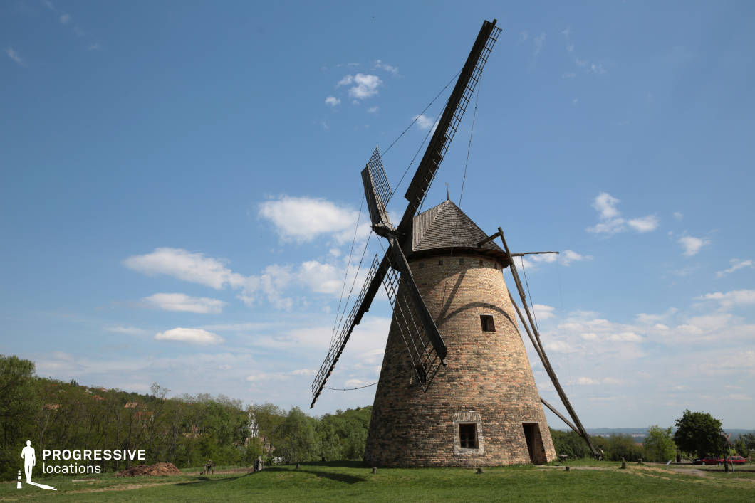 This image captures a striking historic windmill constructed from weathered bricks, standing prominently atop a lush green hill. The structure features a conical roof and large wooden sails, which extend dramatically into the sky, creating a visually compelling silhouette. The surrounding landscape is open and expansive, with distant rolling hills and a tree-lined horizon adding depth and scale to the composition. The rustic charm of the windmill, combined with its well-preserved craftsmanship, makes it an ideal location for period films, historical documentaries, or commercial projects that seek to highlight traditional European rural life.