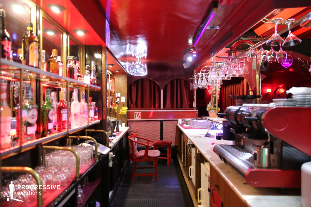 This atmospheric bar features a striking red interior with dim, moody lighting, creating an intimate and slightly vintage ambiance. The narrow space is lined with a well-stocked bar showcasing illuminated liquor bottles on glass shelves, while rows of hanging glasses add depth and texture. A polished counter with classic bar equipment enhances the authentic feel, while velvet drapery and soft, glowing wall sconces contribute to a warm yet slightly mysterious aesthetic. The neon signage and subtle reflections from the glossy ceiling add a modern edge. This location is ideal for filming noir-style dramas, underground club scenes, or late-night rendezvous, offering a visually rich and immersive setting.