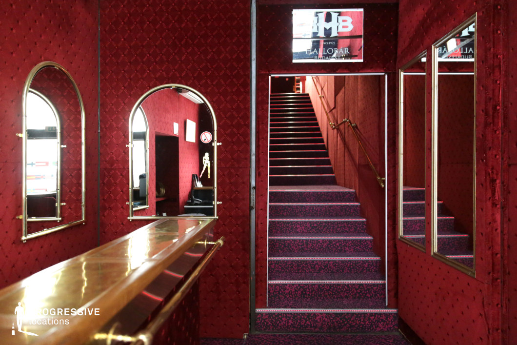 This richly textured interior exudes a vintage, cabaret-like ambiance with deep red velvet walls, gold accents, and a plush patterned carpet leading up a narrow staircase. The arched mirrors and reflective surfaces amplify the depth of the space, while the polished brass bar adds a luxurious yet slightly aged charm. The repetition of gold railings and framed mirrors enhances the sense of symmetry, making the area feel enclosed yet dramatic. This location is perfect for neo-noir films, underground nightlife scenes, or suspenseful thrillers, offering an evocative setting with a blend of decadence and secrecy.