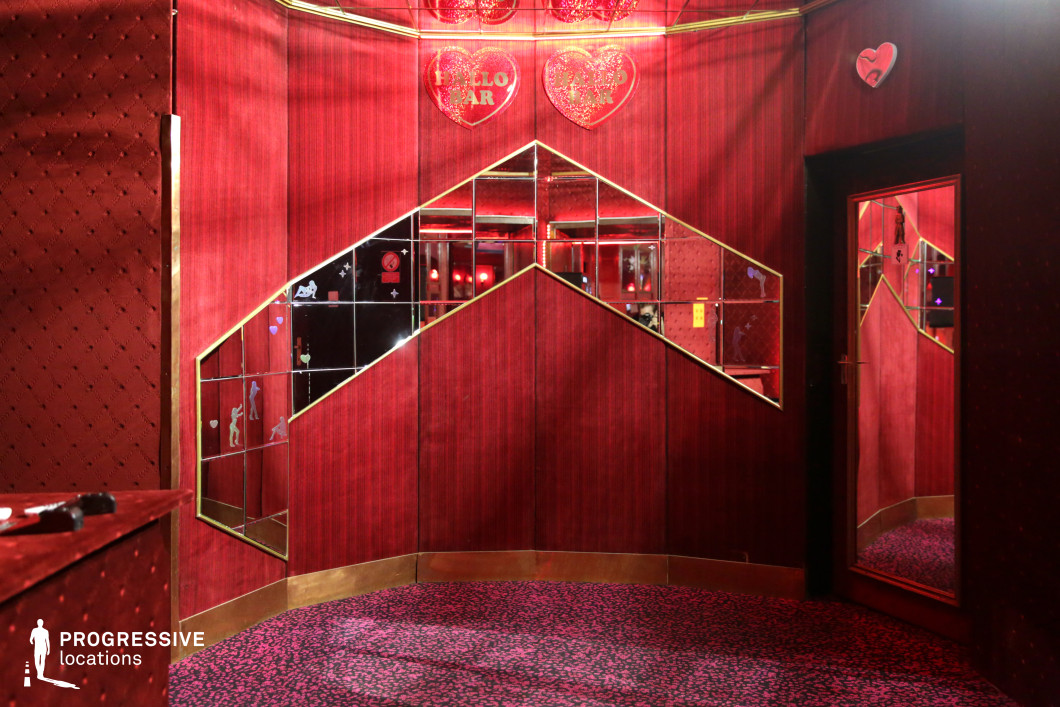 This intimate space is defined by its bold red color palette, featuring plush velvet walls, a patterned carpet, and gold accents that enhance its retro aesthetic. Angular mirrored panels, framed in gold, create a fragmented, almost surreal effect, reflecting the ambient lighting and adding depth to the compact room. Small decorative elements, including heart-shaped signage, reinforce the space’s distinctive, nostalgic character. This location is ideal for neo-noir films, psychological thrillers, or vintage-inspired nightlife scenes, offering an atmospheric setting that blends glamour with an air of mystery.
