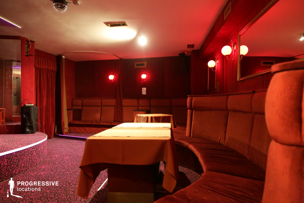 This dimly lit lounge features a retro aesthetic with plush, deep-red upholstered seating arranged in a curved formation around small, draped tables. The walls are lined with dark wood paneling, enhanced by vintage-style wall sconces emitting a warm, reddish glow. A low, circular stage with a subtly illuminated edge serves as the focal point, framed by heavy velvet curtains. The patterned carpet and mirrored wall details add to the nostalgic ambiance, evoking an old-school cabaret or private club setting. This location is ideal for filming noir thrillers, underground crime dramas, or vintage nightclub scenes, offering a rich and moody atmosphere that exudes secrecy and exclusivity.