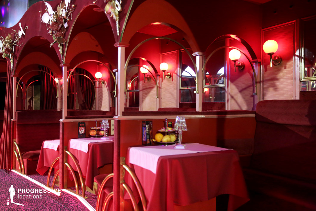 This intimate lounge space features deep red velvet booth seating, arched gold-accented dividers, and soft, ambient lighting from vintage-style wall sconces. The decor blends Art Deco and classic cabaret aesthetics, with rich textures and warm hues creating a seductive, nostalgic atmosphere. The mirrored panels reflect the golden glow of the lamps, enhancing the depth and visual complexity of the space. This setting is ideal for filming noir thrillers, romantic dramas, or underground club scenes, offering a visually striking and immersive backdrop for clandestine meetings, high-stakes conversations, or stylish nightlife sequences.