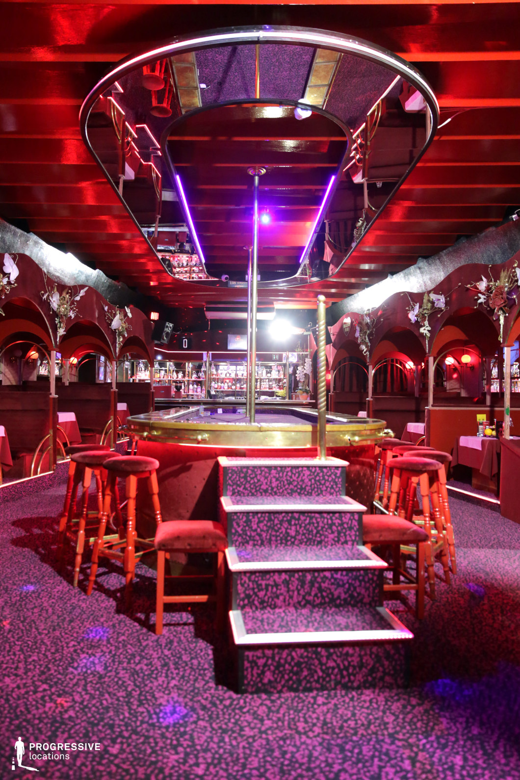 This nightclub features an eye-catching central stage, accessible via carpeted steps, with a mirrored ceiling above reflecting the rich red and purple lighting. The interior design is a mix of classic lounge aesthetics and modern neon accents, with dark wood-paneled walls and arched alcoves creating a sense of depth. The bar in the background is well-stocked and illuminated, adding to the lively atmosphere. Ornate wall decorations and plush seating contribute to an opulent yet intimate ambiance. This setting is ideal for crime thrillers, neo-noir dramas, or high-energy nightlife sequences, providing a dynamic and immersive backdrop for intense confrontations, underground dealings, or glamorous performances.