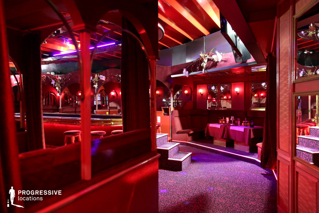 This intimate venue features a rich red color palette, plush seating, and a combination of warm ambient lighting and neon accents, creating a sultry and moody atmosphere. Ornate archways, mirrored surfaces, and dark wood paneling enhance the sense of depth and luxury, while small round tables and booths provide a private, secluded feel. The stage area is subtly integrated, framed by reflective surfaces and decorative floral arrangements, adding to the vintage aesthetic. This location is ideal for noir thrillers, crime dramas, or underground club scenes, offering a visually compelling backdrop for intrigue, secret deals, or nightlife-driven narratives.