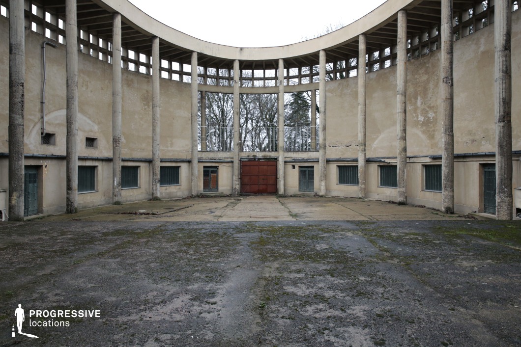 This stark, semi-circular courtyard is defined by weathered concrete columns, a curved facade with small, barred windows, and a rusted metal gate, evoking a sense of decay and forgotten history. The raw, exposed surfaces show signs of aging, with peeling plaster and moss-covered ground contributing to the desolate atmosphere. The repetitive vertical lines of the columns contrast with the enclosed, almost claustrophobic feel of the lower-level windows and doors. This location is ideal for dystopian films, crime dramas, or post-apocalyptic settings, serving as a powerful backdrop for scenes of captivity, clandestine meetings, or abandoned institutional structures.