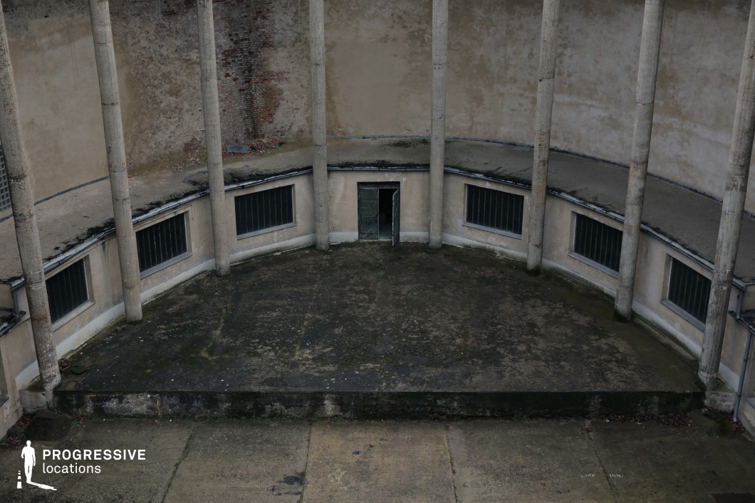 This desolate, circular courtyard is framed by tall, weathered concrete columns and a curved, decayed wall, evoking a stark and oppressive atmosphere. The dark, barred windows and a single open doorway create a sense of mystery and unease. The worn-out concrete flooring, stained with time and exposure, enhances the abandoned aesthetic. The structure’s raw, brutalist design and enclosed form contribute to its cinematic potential. This location is ideal for filming dystopian narratives, psychological thrillers, or prison and interrogation scenes, offering a haunting and isolated backdrop with a strong visual impact.