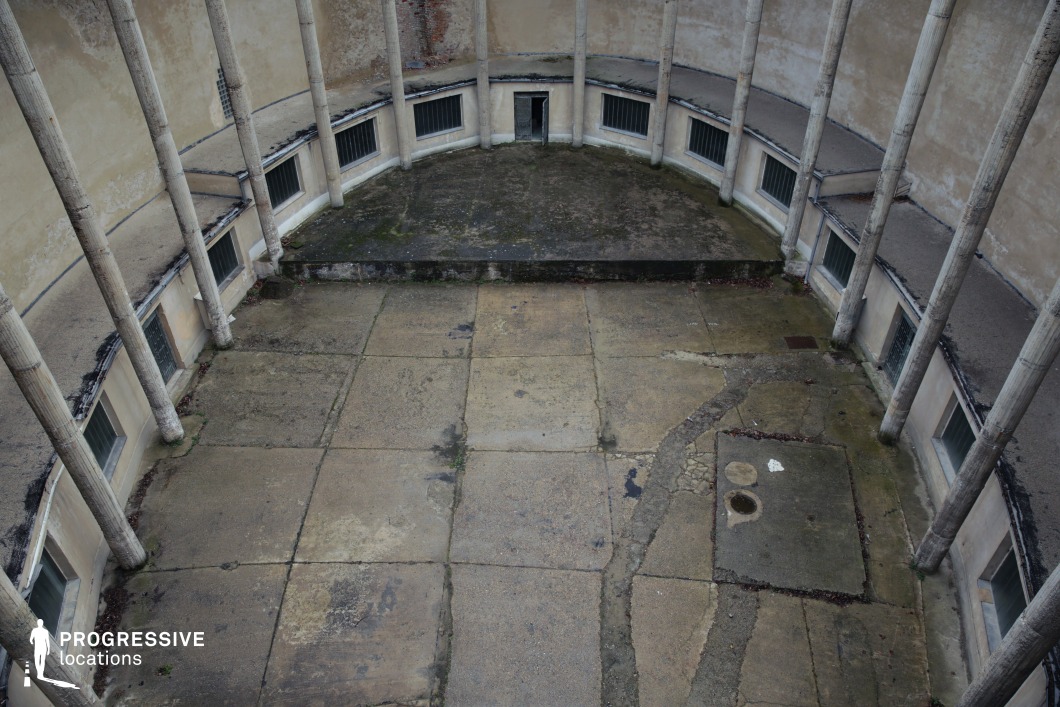 This hauntingly atmospheric space features a large, circular courtyard enclosed by tall, aged columns and worn concrete walls. The cracked and uneven flooring, along with moss-covered surfaces, emphasizes the passage of time and decay. Narrow, barred windows set low into the structure contribute to a confined and isolated feel, while a single, darkened doorway at the far end enhances the sense of mystery. The stark symmetry of the architecture adds to its eerie aesthetic, making this location ideal for psychological thrillers, dystopian films, or historical dramas involving imprisonment, clandestine meetings, or hidden pasts.