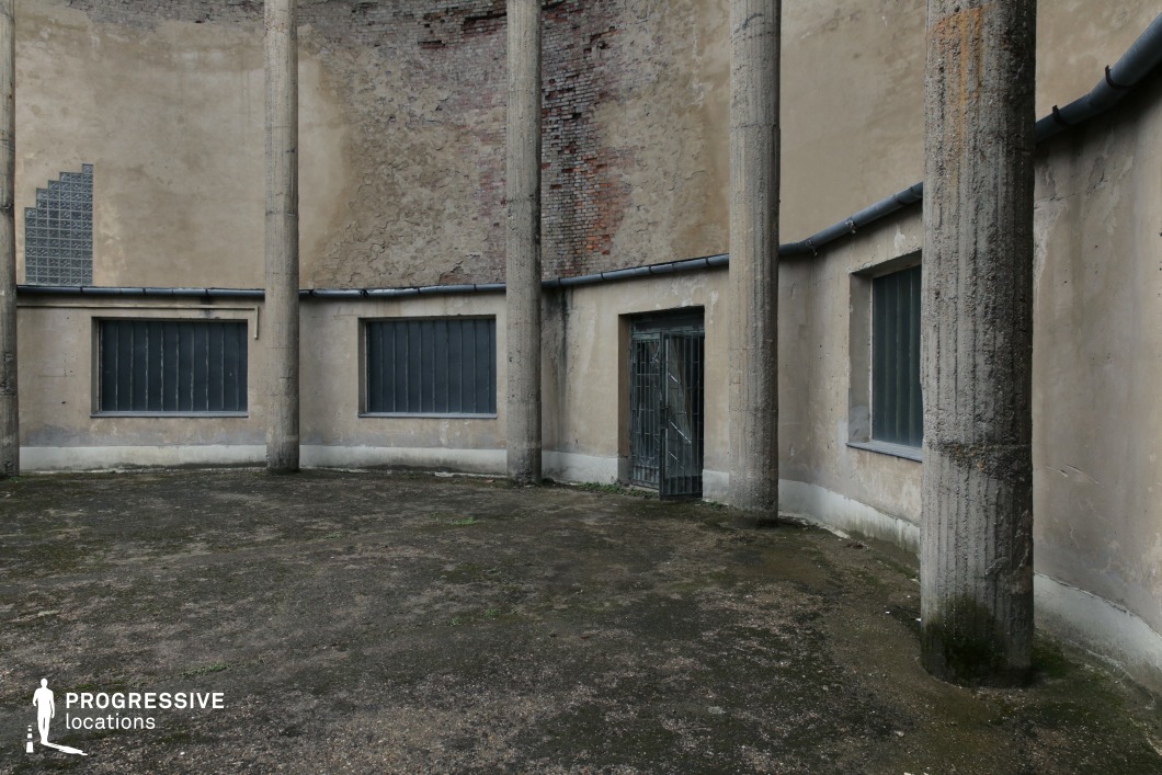 This stark, enclosed courtyard features weathered concrete columns, barred windows, and a rusted metal gate, evoking a sense of decay and isolation. The peeling plaster reveals underlying brickwork, adding texture and history to the setting. The ground is covered in worn, uneven pavement, enhancing the abandoned, industrial feel. The space is defined by harsh geometric lines and a muted color palette, making it an ideal location for dystopian films, crime dramas, or psychological thrillers, offering a visually gritty and atmospheric backdrop with a strong sense of confinement and mystery.