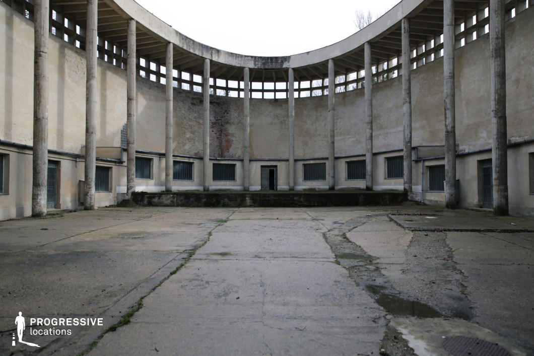 This stark, semi-circular courtyard exudes a sense of desolation and faded grandeur. Tall, weathered concrete columns support a curved open-air gallery, casting long vertical lines that emphasize the structure’s imposing symmetry. The cracked pavement and peeling walls, with remnants of exposed brick, suggest a once-prominent space now left to decay. Small, barred windows and a single central doorway add an air of mystery and isolation. This location is ideal for filming dystopian narratives, psychological thrillers, or historical dramas, offering a raw, atmospheric backdrop that conveys tension, abandonment, and architectural Brutalism.