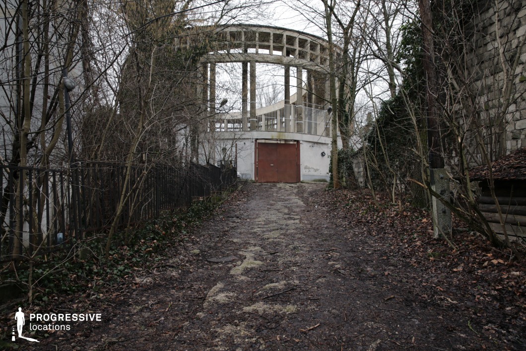 This eerie setting features a weathered, overgrown pathway leading to a decayed modernist structure with an open circular colonnade. The rusted metal doors and stained concrete walls hint at abandonment, while the surrounding leafless trees and creeping ivy enhance the sense of isolation. The uneven ground, enclosed by a mix of iron fencing and stone walls, adds to the unsettling atmosphere. The stark contrast between architectural symmetry and natural decay makes this location ideal for filming psychological thrillers, post-apocalyptic scenes, or mysterious investigative sequences, offering a visually striking and ominous backdrop.