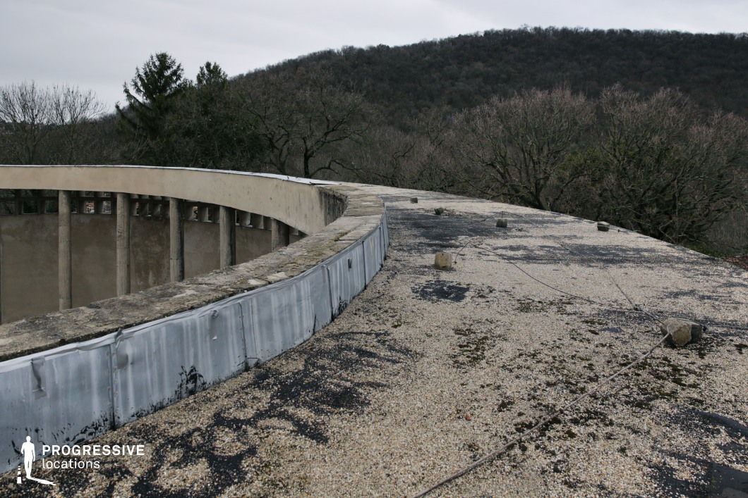 This is a weathered rooftop of an abandoned circular Bauhaus structure. The rooftop is covered in gravel and patches of blackened tar. There is an industrial, aged, abandoned feel. In the background, leafless trees frame rolling forested hills, creating a stark contrast between the built and natural environments. This location is ideal for filming post-apocalyptic scenes, urban exploration narratives, or clandestine meetings, offering a raw and atmospheric setting that evokes isolation and intrigue.