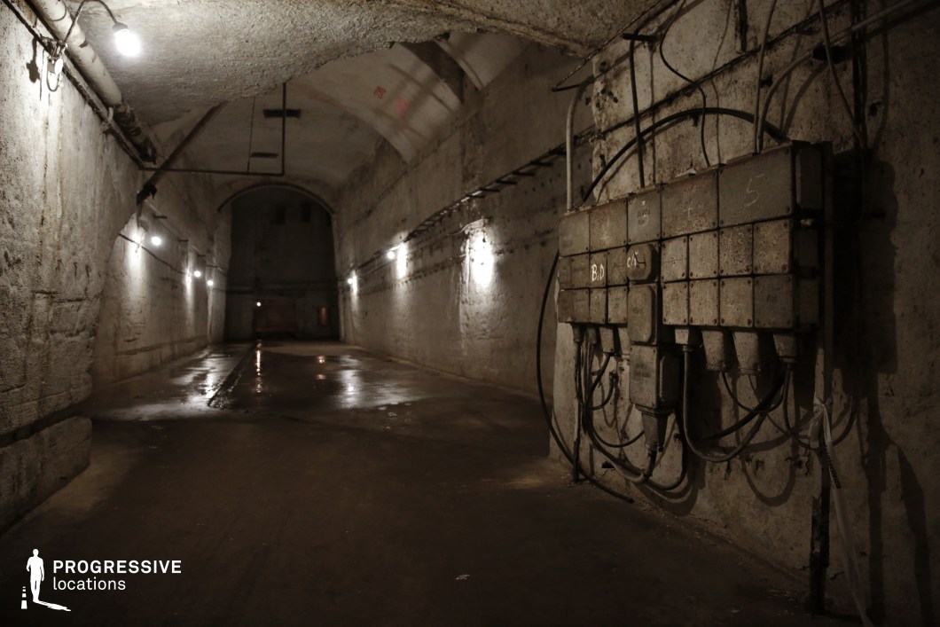 This dimly lit underground tunnel exudes an eerie and industrial atmosphere, with aged concrete walls, exposed wiring, and rusted electrical panels. The arched ceiling, lined with flickering lights, creates a sense of depth and mystery, while damp patches on the floor add to the feeling of neglect. The corroded metal fixtures and scattered pipes contribute to the raw, utilitarian aesthetic. This location is ideal for filming dystopian thrillers, post-apocalyptic settings, or suspenseful escape sequences, providing an immersive and unsettling environment perfect for high-tension storytelling.