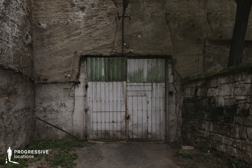 This imposing, weathered metal gate is embedded within a rough-hewn rock wall, giving the location a mysterious and industrial feel. The faded green and white paint, rusted hinges, and makeshift repairs suggest long-term disuse, while the surrounding cracked stone and exposed wiring add to the sense of decay. The uneven cobblestone ground and creeping vegetation enhance the atmosphere of neglect and secrecy. This setting is ideal for thriller, crime, or post-apocalyptic films, serving as an entry point to an underground facility, secret hideout, or forgotten industrial site.