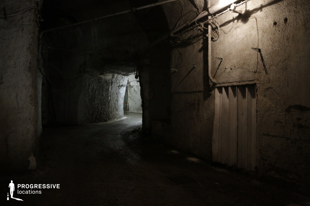 This dimly lit underground tunnel features rugged stone walls and a winding pathway disappearing into the darkness. Exposed wiring, industrial piping, and a weathered wooden door add to the eerie, abandoned atmosphere. The uneven texture of the rock and the sparse artificial lighting create dramatic shadows, enhancing the sense of mystery and isolation. The confined space and rough, utilitarian design make this an ideal filming location for horror, thriller, or post-apocalyptic scenes, lending itself to suspenseful chase sequences, secret hideouts, or the exploration of an abandoned mine or subterranean bunker.