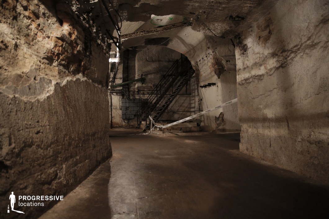 This eerie underground passage features rough-hewn stone walls, exposed brickwork, and scattered remnants of old infrastructure, creating a raw and unsettling atmosphere. The dimly lit space is lined with metal staircases, old electrical panels, and crumbling archways, suggesting a forgotten or abandoned industrial site. The worn floor and makeshift barriers hint at restricted access, adding an element of secrecy. The rugged textures and deep shadows enhance the sense of mystery. This location is ideal for filming horror, thriller, or post-apocalyptic scenes, offering a setting filled with tension, isolation, and a haunting subterranean aesthetic.