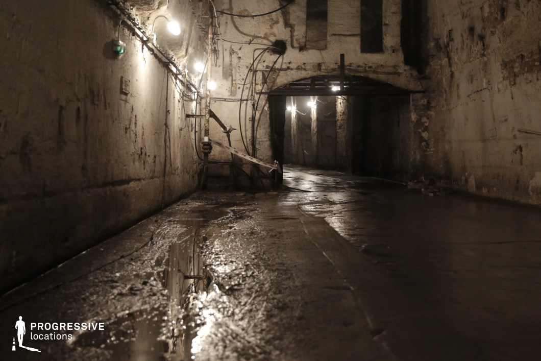 This ominous underground tunnel features damp, stained concrete walls lined with exposed wiring and rusted pipes, creating an atmosphere of decay and secrecy. Dim, flickering lights cast long shadows along the wet, uneven floor, where puddles reflect the sparse illumination. The low arched passageway in the background disappears into darkness, heightening the sense of mystery and unease. The worn industrial textures and abandoned feel make this location ideal for horror films, crime thrillers, or dystopian settings, serving as a backdrop for escape sequences, covert operations, or unsettling discoveries.