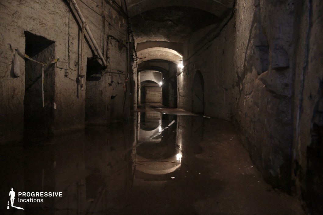 This eerie underground corridor features rough, aged stone walls, exposed pipes, and a vaulted ceiling, creating an unsettling and abandoned atmosphere. Pools of water on the uneven floor reflect the dim artificial lighting, amplifying the sense of decay and isolation. The narrow passageway leads into darkness, with multiple arched doorways and tangled cables adding to the visual complexity. This location is ideal for horror, thriller, or mystery films, providing a claustrophobic and foreboding setting perfect for tension-building scenes or hidden secrets within an urban underworld.