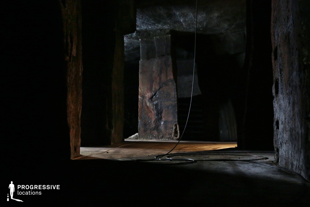 This shadowy, cavernous space features massive, rough-hewn pillars and textured stone or concrete walls, evoking a subterranean industrial or abandoned mining atmosphere. The dim lighting, punctuated by a few streaks of illumination, enhances the eerie, foreboding quality of the scene. Stray cables and scattered debris add to the sense of decay and mystery. The setting is ideal for filming suspenseful, dystopian, or horror narratives, serving as an underground lair, secret hideout, or a perilous exploration site in a post-apocalyptic or sci-fi thriller.