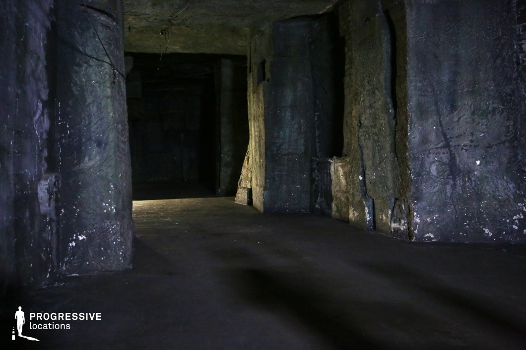 This subterranean space is defined by its rough, hewn stone walls and deep shadows, creating a cavernous and foreboding atmosphere. The uneven textures of the walls, coupled with faint lighting filtering in from an unseen source, enhance the feeling of mystery and isolation. The large, irregular stone pillars contribute to the sense of weight and permanence, while the dark recesses suggest hidden passages or unknown depths. The space is ideal for filming horror, thriller, or fantasy scenes, serving as an ancient underground hideout, an abandoned bunker, or a lost cave system. Its raw, unpolished surfaces and lack of modern elements make it a versatile setting for stories involving secrecy, danger, or supernatural discovery.