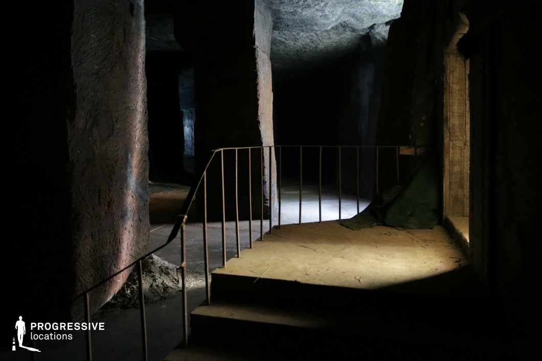 The image showcases a dimly lit underground space with rough, hewn stone walls and a series of shadowed passageways. A simple metal railing lines the edge of a small staircase, which leads into deeper, unilluminated sections of the cavern. The ceiling appears irregular and textured, adding to the raw, natural aesthetic of the space. Selective lighting creates dramatic contrasts between the illuminated areas and the dark recesses, enhancing the sense of mystery and depth. This location is ideal for filming suspenseful, horror, or adventure scenes, well-suited for settings like ancient catacombs, abandoned mines, or hidden underground hideouts in thriller or fantasy narratives.