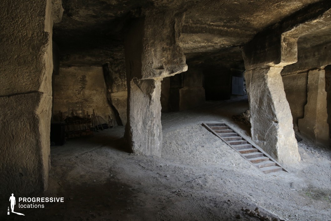 This image depicts a vast underground space carved from solid rock, supported by thick, irregular stone columns that give it a raw, unrefined appearance. The uneven ground, strewn with dust and debris, enhances the rugged feel, while a worn wooden stair structure embedded in the dirt suggests past industrial or mining activity. The dim lighting and shadowy recesses add depth and an element of mystery, making this location an excellent choice for filming adventure, fantasy, or historical dramas. Its cavernous atmosphere and rough-hewn textures also make it suitable for eerie horror sequences, hidden lairs, or archaeological exploration settings.