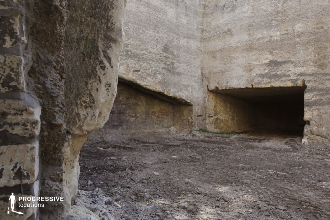 This rugged, excavated quarry features towering stone walls with sharp, uneven cuts, creating a raw and imposing atmosphere. The eroded rock surfaces display a mix of textures and colors, ranging from pale beige to darker earth tones. Two large, rectangular recesses carved into the stone add depth and mystery, resembling ancient dwellings or industrial excavation remnants. The rough, dirt-covered ground enhances the barren and untouched feel of the location. This setting is ideal for filming historical, post-apocalyptic, or fantasy scenes, providing a versatile backdrop for underground hideouts, lost civilizations, or clandestine operations.