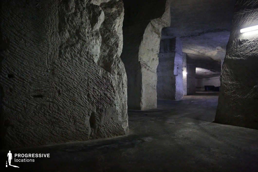 This image depicts a vast underground space with rough-hewn stone walls and massive carved pillars, suggesting an abandoned quarry or cave system. The textured rock surfaces display visible tool marks, adding an industrial and historic character to the setting. The artificial overhead lighting casts a stark contrast between illuminated areas and deep shadows, enhancing the depth and creating a moody, almost ominous atmosphere. The open layout with winding pathways and irregularly shaped support structures makes this location ideal for filming suspenseful, adventure, or sci-fi scenes, such as underground hideouts, secret tunnels, or post-apocalyptic environments.