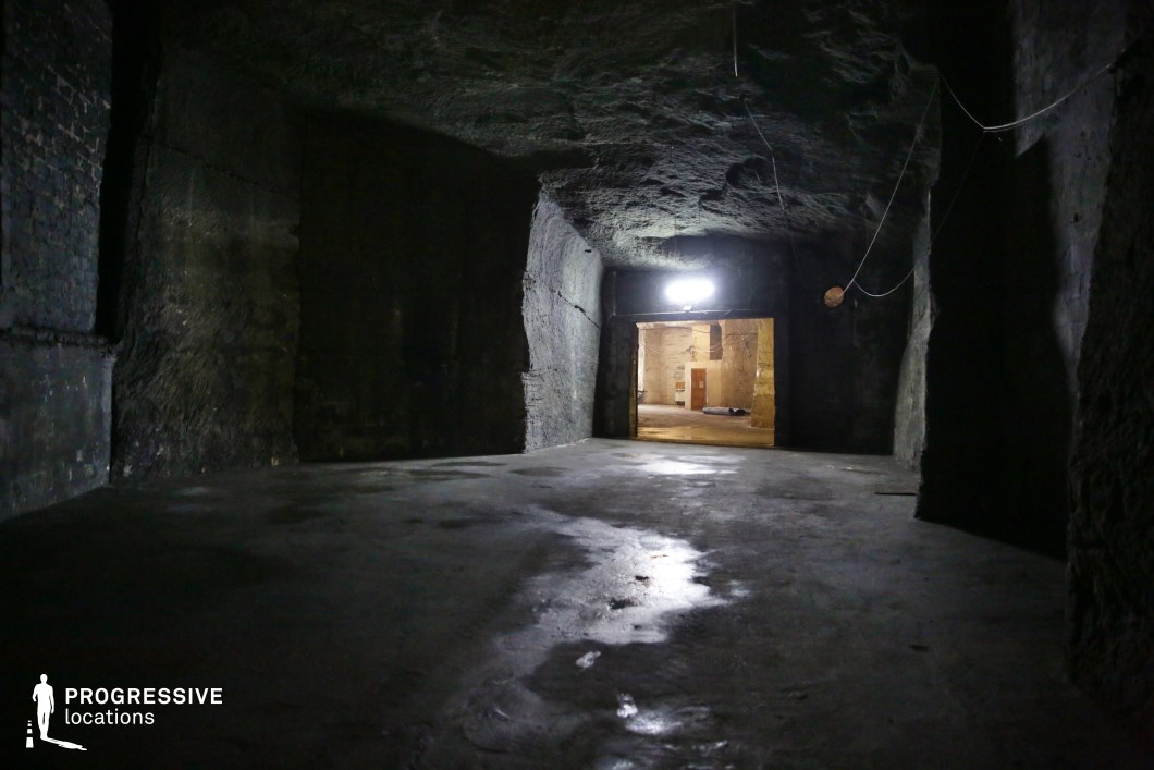 This vast, dimly lit underground chamber features rough-hewn stone walls and a smooth, reflective concrete floor, creating a stark contrast between raw and industrial elements. The uneven ceiling and exposed wiring add to the cavernous, unfinished atmosphere, while the doorway leading to a more illuminated space introduces a sense of depth and mystery. The setting exudes an eerie, secretive quality, making it an ideal location for filming horror, thriller, or post-apocalyptic scenes. It is well-suited for underground hideouts, secret lairs, prison escape sequences, or clandestine meetings in a forgotten subterranean world.