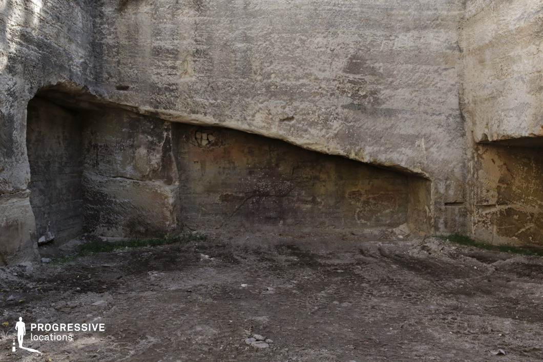 The image captures a weathered stone quarry with rough, textured rock walls enclosing a compact, excavated space. The uneven surfaces and eroded formations suggest a long history of natural and human impact, with visible cracks and discolorations adding depth to the scene. The asymmetrical overhangs create a sense of enclosure, while the raw, earthen floor enhances the location’s rugged and ancient appearance. This setting is ideal for filming historical, fantasy, or post-apocalyptic scenes, offering a primitive and isolated atmosphere suited for survival narratives, hidden sanctuaries, or secret meeting points.