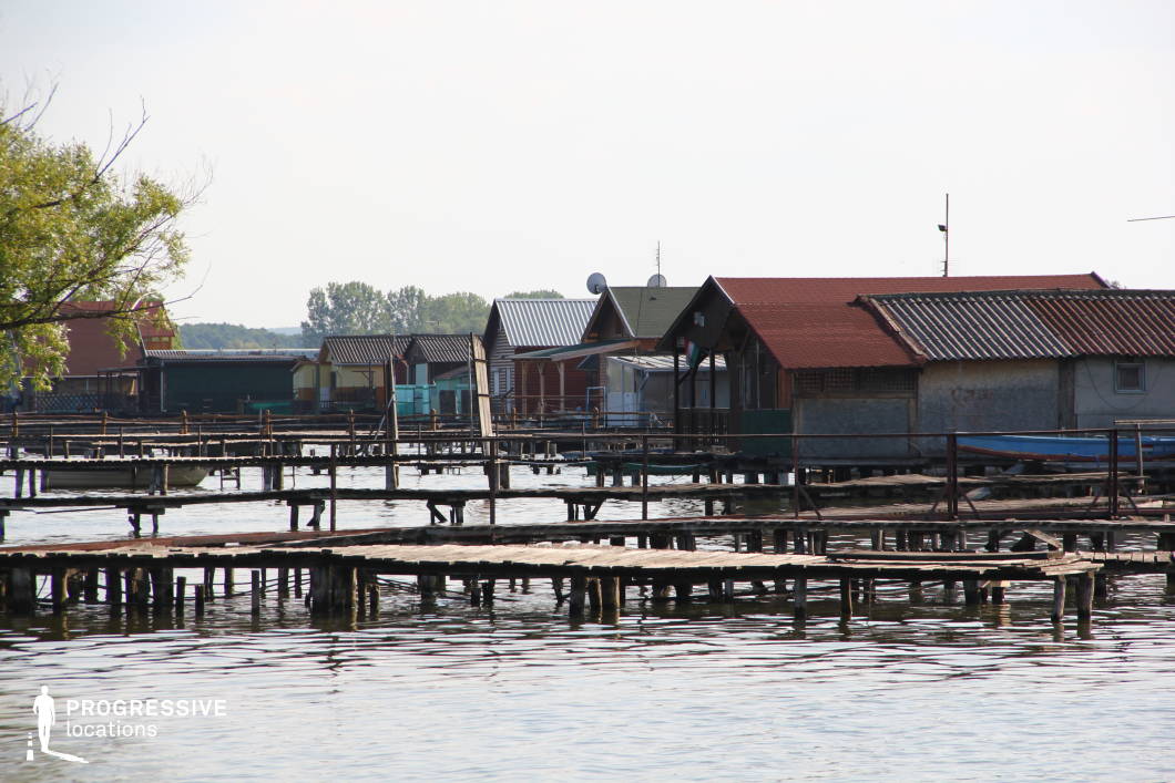 This image captures a unique waterfront settlement composed of rustic fishing huts built on stilts over the water, interconnected by an intricate network of weathered wooden docks. The structures exhibit a mix of metal and wooden roofs, with various colors and materials creating a visually diverse yet cohesive setting. The calm water reflects the aged walkways, adding depth and texture to the scene. The location exudes a quiet, remote atmosphere, making it ideal for filming independent dramas, contemplative character studies, or mystery and thriller narratives that require an isolated, off-the-grid environment.