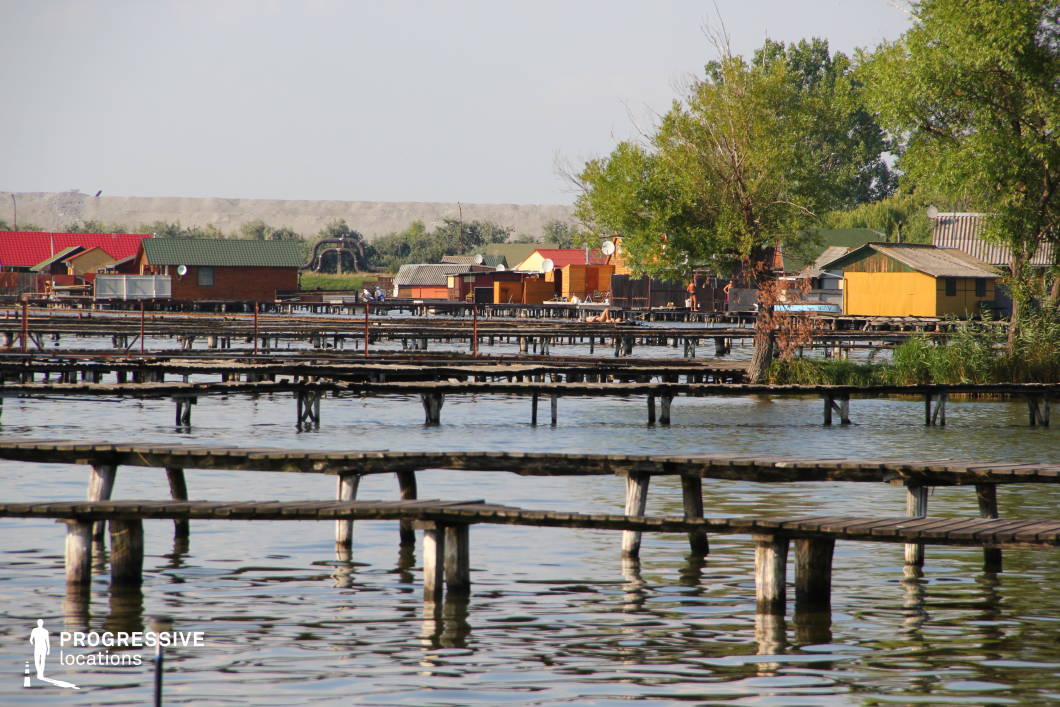 This lakeside settlement features an intricate network of narrow wooden piers stretching across calm waters, leading to a collection of colorful, rustic houses on stilts. The reflections of the walkways on the water create a layered visual effect, enhancing the sense of depth and perspective. The diverse architectural styles, with vibrant rooftops and wooden facades, contrast against the natural greenery along the shoreline. The combination of water, wooden structures, and open space makes this location ideal for filming tranquil rural scenes, atmospheric mystery sequences, or character-driven dramas set in a secluded lakeside community.