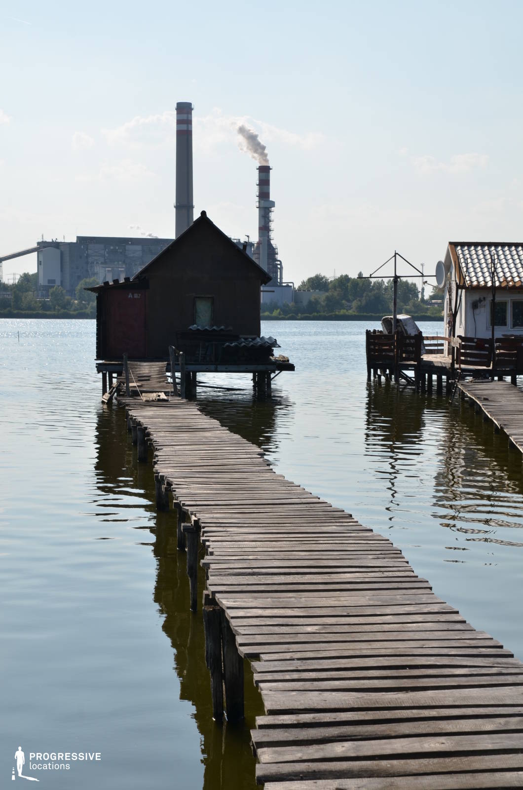 This image presents a contrast between a rustic waterside settlement and an imposing industrial complex in the background. The foreground features a weathered wooden pier extending across the calm water towards a small, simple house on stilts, with uneven planks enhancing the raw, handmade aesthetic. Additional wooden structures line the water’s edge, emphasizing a sense of isolation and resilience. In stark contrast, the background is dominated by towering smokestacks and large industrial buildings, suggesting themes of environmental tension or socioeconomic disparity. This setting is ideal for filming introspective dramas, dystopian thrillers, or crime narratives, offering a visually compelling blend of rural simplicity and industrial dominance.