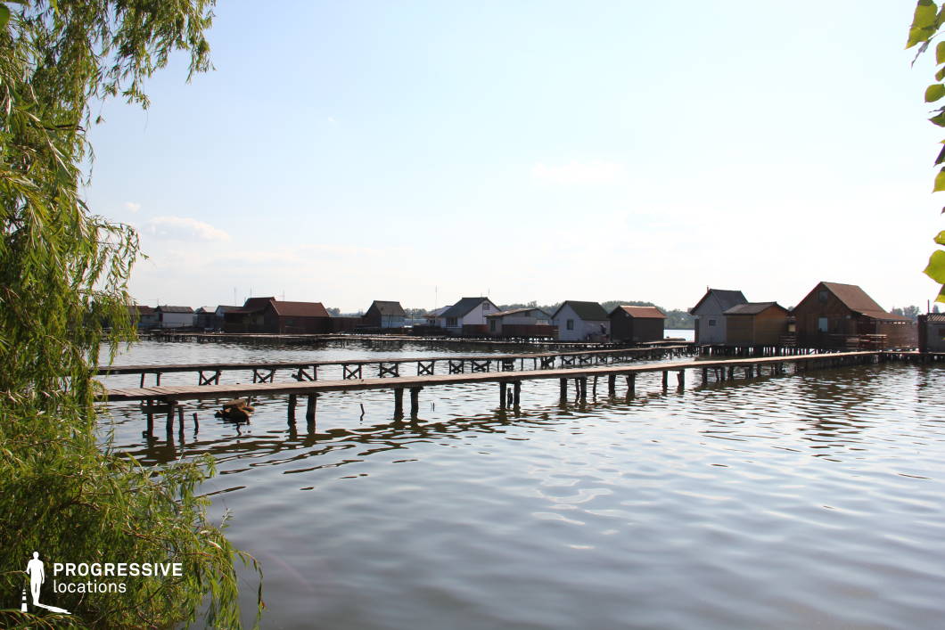 This serene lakeside scene features a row of wooden huts built on stilts above the water, connected by a network of rustic wooden piers. The simple, functional architecture of the huts, with slanted roofs and neutral tones, blends harmoniously with the natural surroundings. The gently rippling water reflects the sky, adding a sense of calm and isolation. Overhanging branches in the foreground frame the setting, enhancing its secluded and picturesque atmosphere. This location is ideal for filming tranquil, introspective, or atmospheric scenes, making it perfect for dramas, romantic narratives, or mystery films set in a remote fishing village or an off-the-grid retreat.