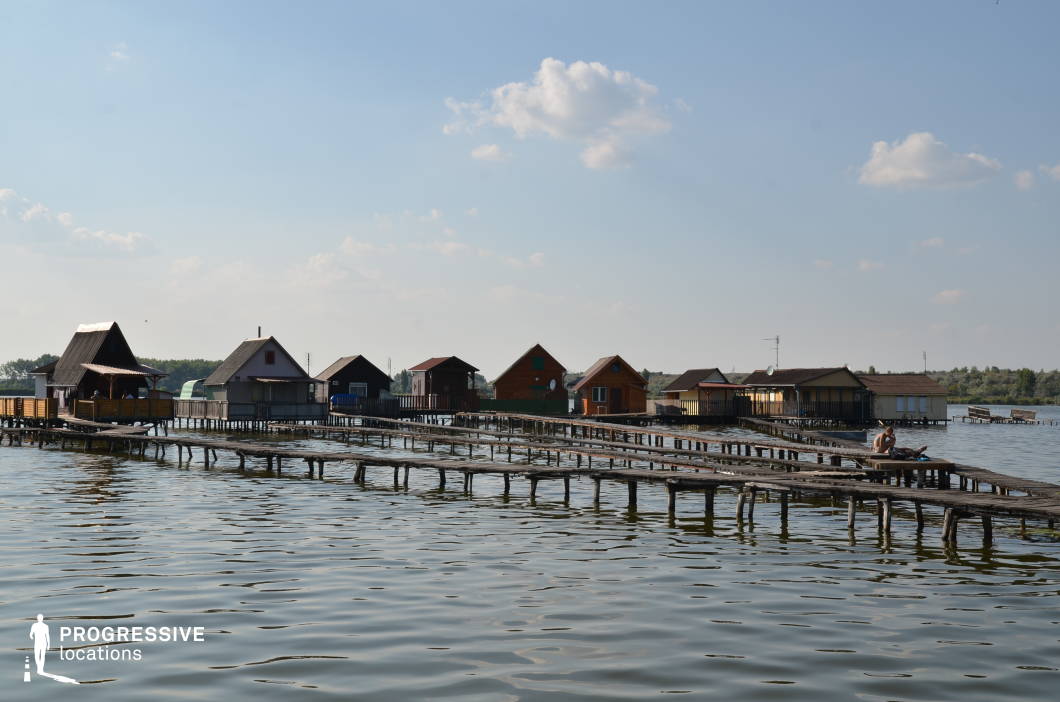 The image presents a picturesque lakeside settlement with small wooden houses built on stilts above the water, connected by narrow, weathered wooden walkways. The structures vary in color and design, contributing to a rustic and serene atmosphere. The calm water reflects the houses and the sky, enhancing the tranquil setting. The distant shoreline, lined with trees and vegetation, provides a natural backdrop. This location is ideal for filming atmospheric dramas, independent films, or mystery stories, as well as travel documentaries capturing off-the-grid lifestyles or unique waterside communities.