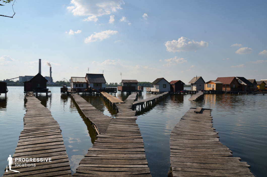 This unique lakeside settlement features a network of weathered wooden boardwalks extending over calm waters, leading to small stilted houses in various muted and earthy tones. The rustic construction and irregular paths create an isolated, almost surreal atmosphere, while the distant industrial chimneys contrast the tranquil setting with an imposing urban presence. The visual juxtaposition between the natural lake environment and the industrial background adds depth and complexity. This location is ideal for filming mystery, indie drama, or dystopian stories, lending itself well to narratives about isolation, hidden communities, or contrasting worlds of industry and nature.