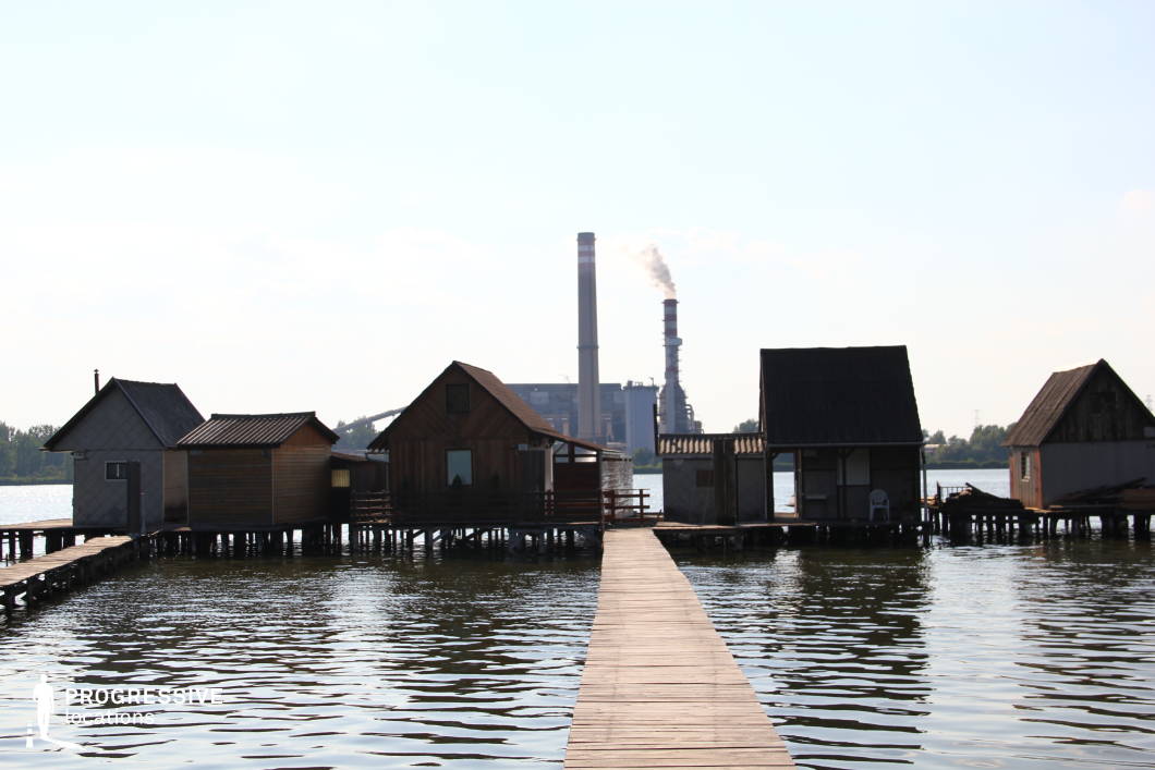 This unique lakeside setting features wooden stilt houses connected by narrow boardwalks, creating a rustic and isolated atmosphere. The weathered wooden structures contrast starkly with the looming industrial power plant in the background, where tall smokestacks rise above the horizon. The still water reflects both the huts and the factory, enhancing the visual tension between the natural and the industrial. This location is ideal for filming atmospheric dramas, dystopian thrillers, or social commentaries, providing a striking setting for stories exploring isolation, environmental themes, or the clash between tradition and modern industry.