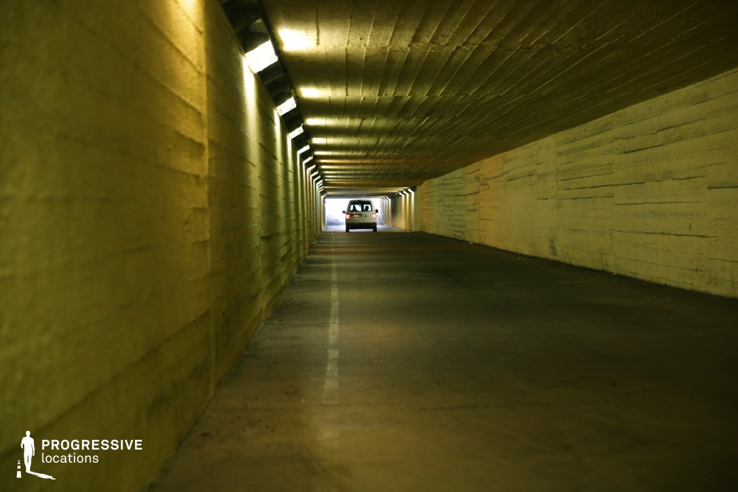 This long, narrow concrete underpass extends into the distance, illuminated by a row of overhead lights that cast a yellowish glow on the textured walls. The rough, unfinished concrete and the low ceiling create a confined, slightly oppressive atmosphere, while the vanishing point at the tunnel’s exit adds a sense of depth and movement. The play of artificial lighting and shadows enhances the space’s gritty, utilitarian aesthetic. This setting is ideal for filming tense chase sequences, urban crime dramas, or psychological thrillers, as it conveys a sense of isolation, secrecy, or impending action. The underpass could also serve as a transitionary space in narratives involving escape, confrontation, or clandestine activity.