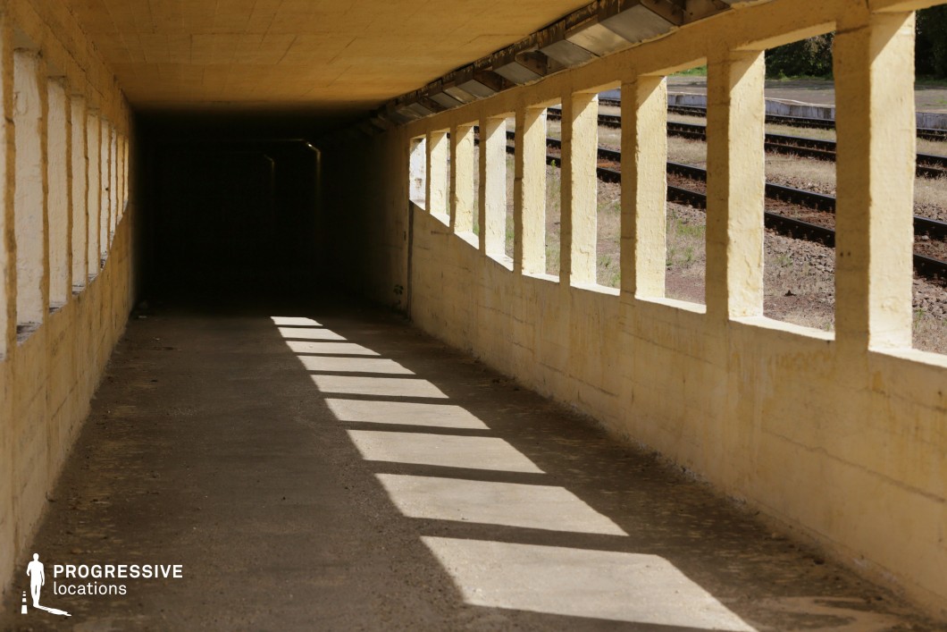 This aged pedestrian underpass, constructed from yellowed concrete, features a series of rectangular openings along one side, allowing glimpses of railway tracks and casting sharp, rhythmic shadows on the ground. The passage narrows into darkness, creating a visually striking contrast between the bright exterior and the ominous interior. The rough, unpolished surfaces and lack of modern infrastructure suggest abandonment or minimal upkeep. This location is perfect for filming tense, suspenseful scenes in thrillers, noir films, or dystopian settings, offering a confined space ideal for chase sequences, dramatic confrontations, or psychological tension.