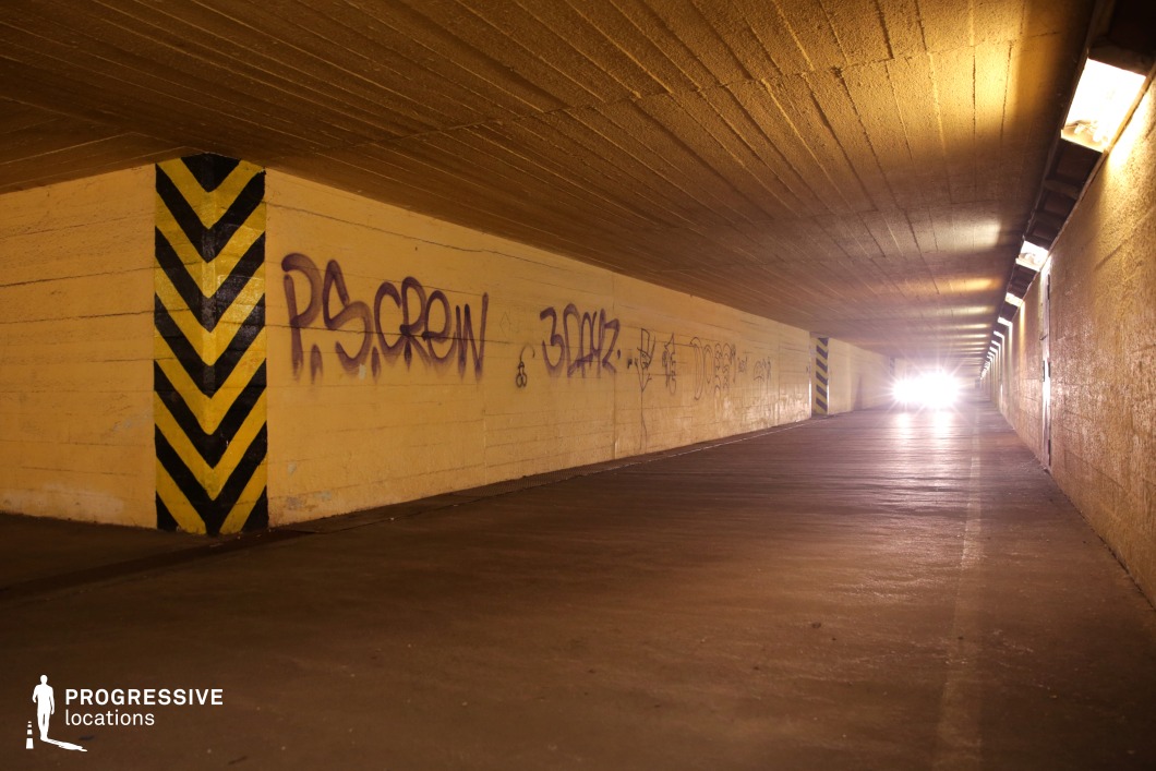 The image depicts a dimly lit concrete underpass with worn yellow walls covered in graffiti, creating an urban and raw aesthetic. The tunnel's low ceiling and repeating overhead lights emphasize its confined nature, while the bold black and yellow hazard markings add a sense of caution. The rough texture of the walls and floor enhances the location's gritty atmosphere. A vehicle's bright headlights in the distance introduce a dramatic focal point, adding depth and tension. This setting is ideal for filming crime, neo-noir, or thriller scenes, particularly chase sequences, secretive meetings, or tense confrontations in an underground, urban environment.